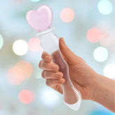 A hand holds the Gläs 8" Sweetheart Glass Dildo from Gläs, crafted from hypoallergenic material. It showcases a transparent, wavy rod topped with a pink heart-shaped stopper. The softly blurred background is decorated with circular lights in an array of colors.